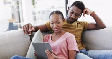 Mutlu Afro-Amerikan çifti tablet kullanıyor ve evdeki koltukta yavaş çekimde konuşuyorlar. Yaşam tarzı, ilişki, birliktelik, rahatlama, iletişim ve ev hayatı, değişmemiş..