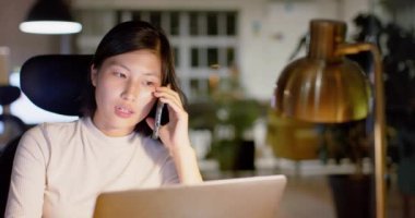 Asyalı sıradan bir iş kadını akıllı telefondan konuşur ve gece ofiste dizüstü bilgisayar kullanır, ağır çekimde. İş, iletişim, günlük ofis, teslim tarihi ve geç saatlere kadar çalışma, değiştirilmemiş.