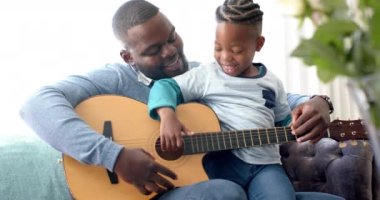 Mutlu Afro-Amerikan baba ve oğlu kanepede oturup gitar çalıyor, ağır çekimde. Müzik, aile, yaratıcılık, öğrenme, keşif ve birliktelik, değiştirilmemiş.