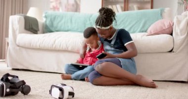 Afrika kökenli Amerikalı kız ve erkek kardeşler oturma odasında akıllı telefon ve tablet kullanıyorlar, ağır çekimde. Aile, iletişim, çocukluk, aile hayatı, değişmemiş.