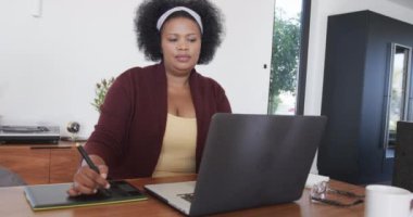 Afro-amerikan kadın diz üstü bilgisayar ve grafik tablet kullanıyor, değiştirilmemiş, ağır çekimde. Yaşam tarzı, beden içeriği, iletişim ve ev hayatı..