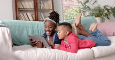 Mutlu Afro-Amerikan kız kardeş kanepeye uzanmış ve tablet kullanıyor, ağır çekimde. Aile, iletişim, çocukluk, yaşam tarzı, bakım ve aile hayatı, değiştirilmemiş.