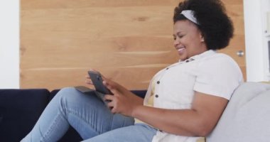 Mutlu Afro-Amerikan artı uzun boylu kadın koltukta oturuyor ve tablet kullanıyor, değiştirilmemiş, ağır çekimde. Yaşam tarzı, beden içeriği, iletişim ve ev hayatı..