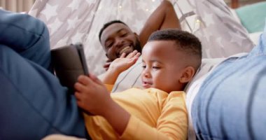 Mutlu Afro-Amerikan baba ve oğlu tablet kullanıyor ve evde yavaş çekimde beşlik çakıyor. Aile, iletişim, babalık, çocukluk, aile hayatı, değişmemiş.