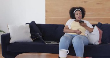 Afro-amerikan kadın koltukta oturuyor ve akıllı telefonla konuşuyor, değiştirilmemiş, ağır çekimde. Yaşam tarzı, beden içeriği, iletişim ve ev hayatı..