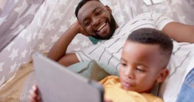 Mutlu Afro-Amerikan baba ve oğlu tablet kullanıyor ve evde gülümsüyor, ağır çekimde. Aile, iletişim, babalık, çocukluk, aile hayatı, değişmemiş.