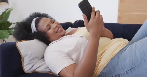 快乐的非洲裔美国人 身材的女人躺在沙发上 用智能手机 毫发无损 动作缓慢 生活方式 身体包容性 沟通和家庭生活 — 图库视频影像