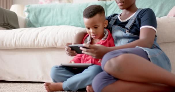 stock video African american sister and brother using tablet and smartphone in living room, slow motion. Family, communication, childhood, domestic life, unaltered.