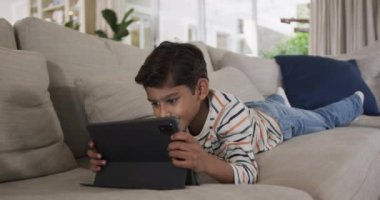 Çift ırklı çocuk kanepeye uzanıp tablet kullanıyor, ağır çekimde. Boş zaman, iletişim, rahatlama ve ev hayatı, değişmemiş..