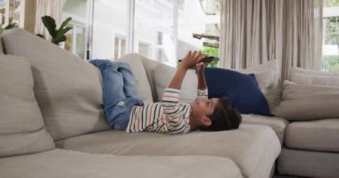 Çift ırklı çocuk kanepeye uzanıp tablet kullanıyor, ağır çekimde. Boş zaman, iletişim, rahatlama ve ev hayatı, değişmemiş..