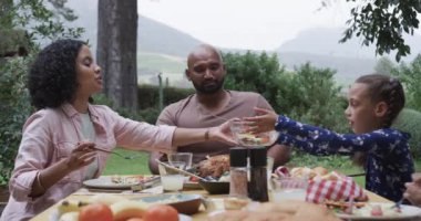 Mutlu çift ırklı ebeveynler, oğlu ve kızı bahçede yemek yiyorlar, ağır çekimde. Aile, yemek, birliktelik, sağlıklı yaşam, yaşam tarzı ve ev hayatı, değişmemiş..