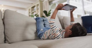 Çift ırklı çocuk kanepeye uzanıp tablet kullanıyor, ağır çekimde. Boş zaman, iletişim, rahatlama ve ev hayatı, değişmemiş..