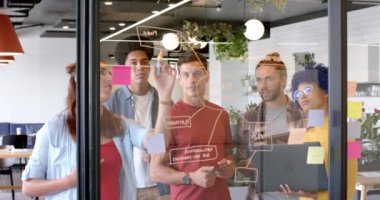 Dizüstü bilgisayarla beyin fırtınası yaparak cam duvara notlar yazarak, yavaş çekimde mutlu yaratıcı meslektaşlar. Günlük ofis, takım çalışması, beyin fırtınası, iletişim, iş ve yaratıcı çalışma, değiştirilmemiş.