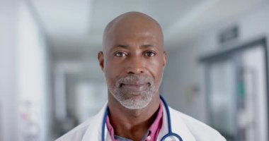 Kameraya bakıp gülümseyen, ağır çekimde mutlu bir Afrikalı Amerikalı erkek doktorun portresi. Hastane, ilaç ve sağlık hizmetleri değiştirilmemiş..