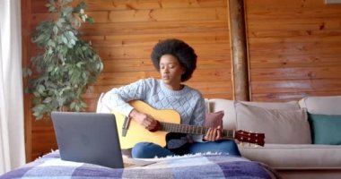 Yavaş çekimde, oturma odasında dizüstü bilgisayar kullanarak gitar çalan Afrikalı Amerikalı kadın. Rahatlama, iletişim, yaşam tarzı ve ev hayatı, değişmemiş..