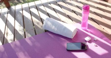 Havlu, su şişesi, akıllı telefon ve kulaklık güneşli terasta yoga minderinde, ağır çekimde. Refah, iletişim, rahatlama, sağlıklı yaşam tarzı ve ev hayatı.