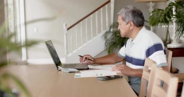 Çift ırklı kıdemli bir adam evrak işleriyle uğraşıyor ve evde dizüstü bilgisayar kullanıyor, ağır çekimde. Finans, emeklilik, ev hayatı ve yaşlı yaşam tarzı, değişmemiş.