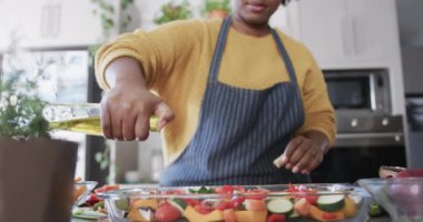 Önlüklü mutlu Afrikalı Amerikalı kadın mutfakta sebzelere yağ döküyor, ağır çekimde. Yemek, yemek, sebze, sağlıklı yaşam, refah ve ev hayatı, değişmemiş.