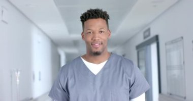 Portrait of happy biracial male doctor wearing scrubs, smiling in corridor, slow motion. Hospital, medicine, healthcare and work, unaltered.