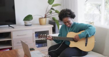 Mutlu Afro-Amerikan kadın gitar çalıyor, evde dizüstü bilgisayar ve kulaklık kullanıyor, ağır çekimde. Hobiler, müzik, çevrimiçi öğrenme, iletişim, rahatlama ve yaşam tarzı, değiştirilmemiş.