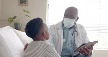 Kıdemli Afro-Amerikan erkek doktor tablet kullanıyor. Evde hasta çocukla konuşuyor. Ağır çekimde. Ev ziyareti, iletişim, hijyen, sağlık hizmetleri, sağlık ve çocukluk, değişmemiş