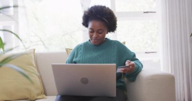 Mutlu Afrikalı Amerikalı kadın evde kredi kartı ve laptop kullanarak ödeme yapıyor, ağır çekimde. Çevrimiçi alışveriş, boş zaman, iletişim, yaşam tarzı ve ev hayatı, değiştirilmemiş.