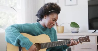 Mutlu Afro-Amerikan kadın gitar çalıp şarkı söylüyor. Evdeki koltukta oturuyor. Ağır çekimde. Hobiler, müzik, yaratıcılık, rahatlama, yaşam tarzı ve ev hayatı, değişmemiş.
