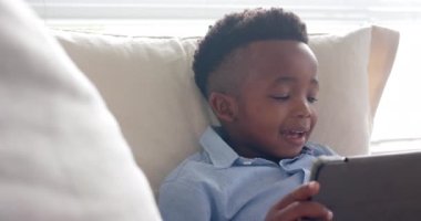 Mutlu Afrikalı Amerikalı çocuk kanepede uzanıp tablet kullanıyor, ağır çekimde. Yaşam tarzı, iletişim, teknoloji, çocukluk ve ev hayatı, rahatlama, değişme.