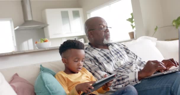 stock video African american grandfather with grandson using tablet and laptop on couch at home, slow motion. Family, lifestyle, childhood, communication, relaxation and domestic life, unaltered.