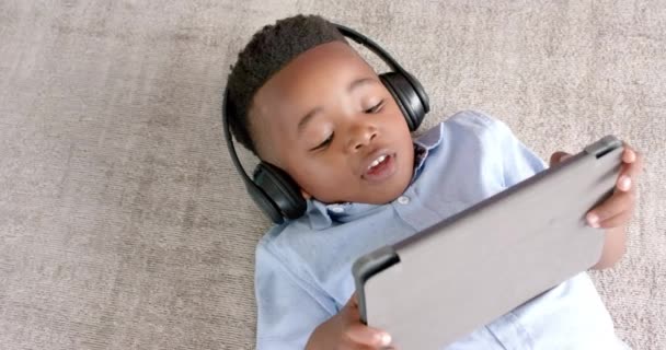 Lykkelig Afroamerikansk Gutt Med Hodetelefoner Liggende Gulvet Med Nettbrett Hjemme – stockvideo