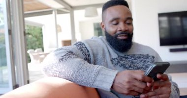 Mutlu Afro-Amerikan adam güneşli oturma odasında akıllı telefon kullanıyor, ağır çekimde. Rahatlama, yaşam tarzı, iletişim ve ev hayatı, değişmemiş..