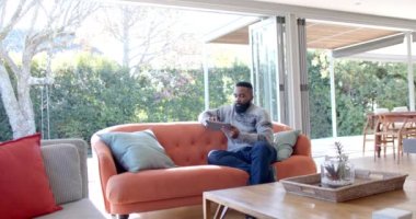 Afro-Amerikalı adam güneşli oturma odasında tablet ve kredi kartı kullanıyor, ağır çekimde. Rahatlama, yaşam tarzı, iletişim ve ev hayatı, değişmemiş..