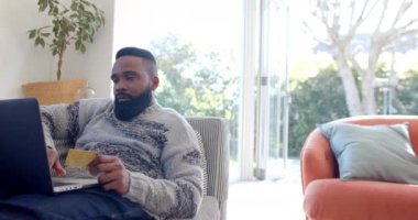 Mutlu Afro-Amerikalı adam güneşli oturma odasında dizüstü bilgisayar ve kredi kartı kullanıyor, ağır çekimde. Rahatlama, yaşam tarzı, iletişim ve ev hayatı, değişmemiş..