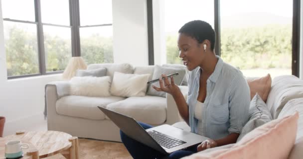 幸せなアフリカ系アメリカ人の女性は ソファに座ってスマートフォンで話していると ラップトップを使用して スローモーション ライフスタイル コミュニケーション 国内生活変更なし — ストック動画