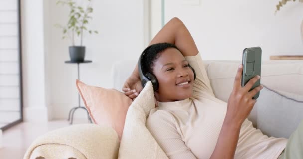 幸せなアフリカ系アメリカ人の女性は ヘッドフォンを着用し スマートフォン スローモーションを使用してソファに横たわっています リラクゼーション ライフスタイル コミュニケーション 国内生活 変わらない — ストック動画