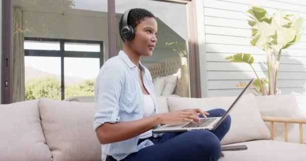 アフリカ系アメリカ人の女性は ラップトップ スローモーションを使用して ヘッドフォンを着てテラスのソファに座っている ライフスタイル コミュニケーション 国内生活変更なし — ストック動画