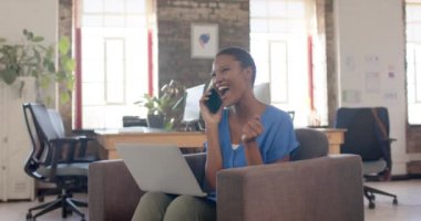 Happy african american casual businesswoman talking on smartphone in creative office, slow motion. Business, technology, celebration and work, unaltered.