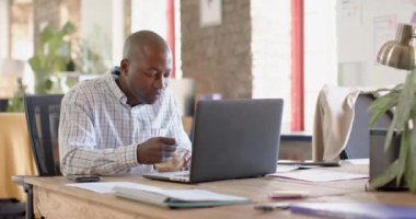 Happy african american casual businessman using smartphone and having lunch in office, slow motion. Business, food, technology and work, unaltered.