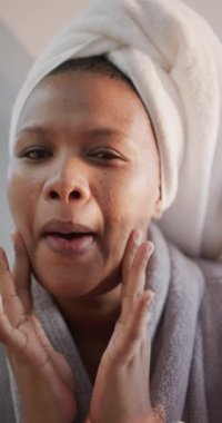 Kafasında havlu olan mutlu Afro-Amerikan kadının banyoya krem sürerken çekilmiş dikey videosu. Kişisel bakım, sağlık ve güzellik, refah ve yaşam tarzı, değişmemiş..