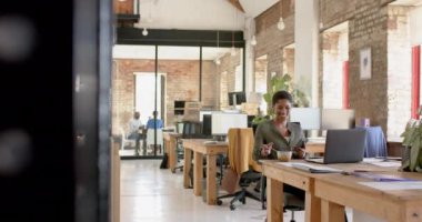 Happy african american casual businesswoman using smartphone and having lunch in office, slow motion. Business, food, technology and work, unaltered.