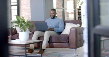 Happy african american casual businessman having laptop video call on couch, copy space, slow motion. Business, work, communication and casual office, unaltered.