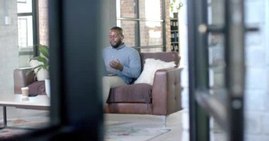 Happy african american casual businessman having laptop video call on couch, copy space, slow motion. Business, work, communication and casual office, unaltered.