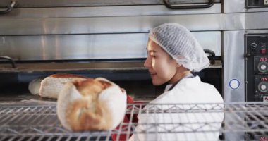 Fırın mutfağında çalışan mutlu Asyalı fırıncı kadın yavaş çekimde taze ekmek çıkarıyor. Yiyecek, küçük işletme, fırın, fırın ve iş, değiştirilmemiş.