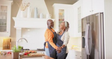Afro-Amerikan önlüklü mutlu çift mutfakta dans ediyor, ağır çekimde. Emeklilik, ev hayatı, yemek pişirme, birliktelik, sağlıklı yaşam ve kıdemli yaşam tarzı, değişmemiş.