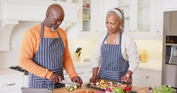 Afrikkalainen Amerikkalainen Vanhempi Pari Esiliinoissa Valmistamassa Ateriaa Käyttäen Tablettia Keittiössä — kuvapankkivideo