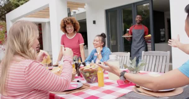 アフリカ系アメリカ人男性は 庭のディナーテーブルで友人の多様なグループに食事を提供しています 食べ物 お祝い トータルネス ライフスタイル 家庭生活 変わらない — ストック動画