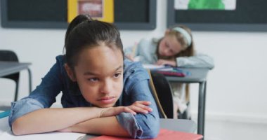 Masada oturan sıkılmış, yorgun, melez bir kızın videosu. Sınıfta kafa üstü uzanıyor, fotokopi çekiyor. Eğitim, çocukluk, kapsayıcılık, ilkokul ve öğrenim kavramı.