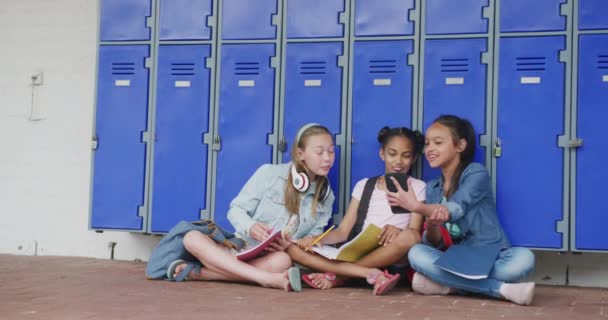 スマートフォンを見ている3人の幸せで多様な女子生徒のビデオ 廊下に座ってコピースペース 幼少期 小学校 学習コンセプト — ストック動画