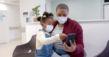 African american mother and daughter wearing face masks using smartphone in hospital, slow motion. Medicine, healthcare services, family, childhood, communication and hospital, unaltered.