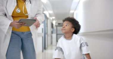 Tabletli Afrikalı Amerikalı kadın doktor koridorda koltuk değneklerini kullanarak kızla yürüyor, ağır çekimde. İlaç, sağlık hizmetleri, çocukluk ve hastane, değiştirilmemiş.